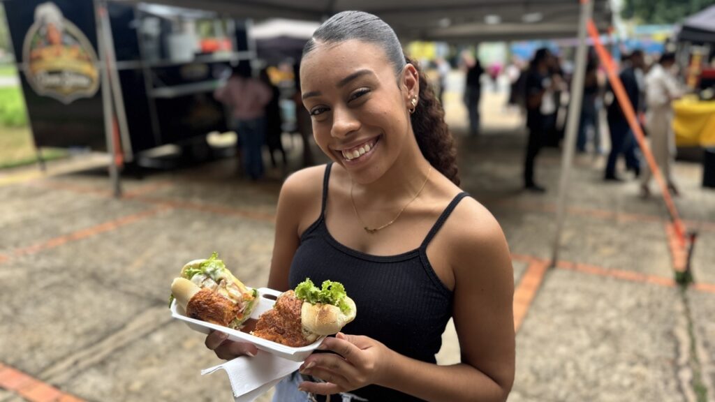 A imagem mostra uma jovem sorridente segurando uma bandeja branca com dois sanduíches grandes e apetitosos. Ela está em um ambiente ao ar livre, possivelmente um mercado ou festival de comida, com barracas e um food truck ao fundo. A jovem tem cabelo escuro preso em um rabo de cavalo, usa uma blusa preta sem mangas, brincos pequenos e um colar delicado. Os sanduíches parecem estar recheados com carne, alface e molho. A foto foi tirada em um momento de luz natural, com foco nítido nela e nos sanduíches, enquanto o fundo está desfocado, destacando a movimentação do local.