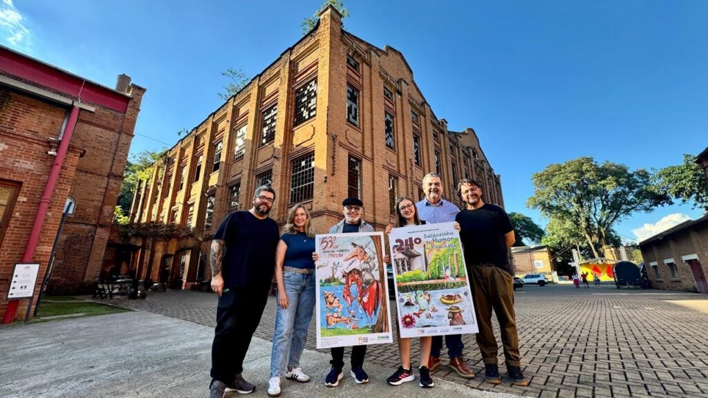 A imagem mostra um grupo de seis pessoas posando para uma foto ao ar livre, em frente a um grande prédio histórico de tijolos, com várias janelas e pequenas árvores crescendo em seu telhado. O grupo é composto por quatro homens e duas mulheres, todos vestidos de forma casual. Dois deles seguram cartazes verticais; um cartaz apresenta uma caricatura de Dom Quixote e o texto "51 Salão Internacional de Humor de Piracicaba", enquanto o outro cartaz tem uma ilustração colorida com o texto "22 Salãozinho de Humor de Piracicaba". O ambiente parece ser uma praça ou pátio pavimentado, com o céu azul e o sol baixo, indicando fim de tarde. Ao fundo, além do prédio principal, há outras construções de tijolos, árvores e uma estrutura que parece ser um palco ou tenda. A foto foi tirada de um ângulo baixo, o que valoriza a altura do prédio e destaca as pessoas em primeiro plano.