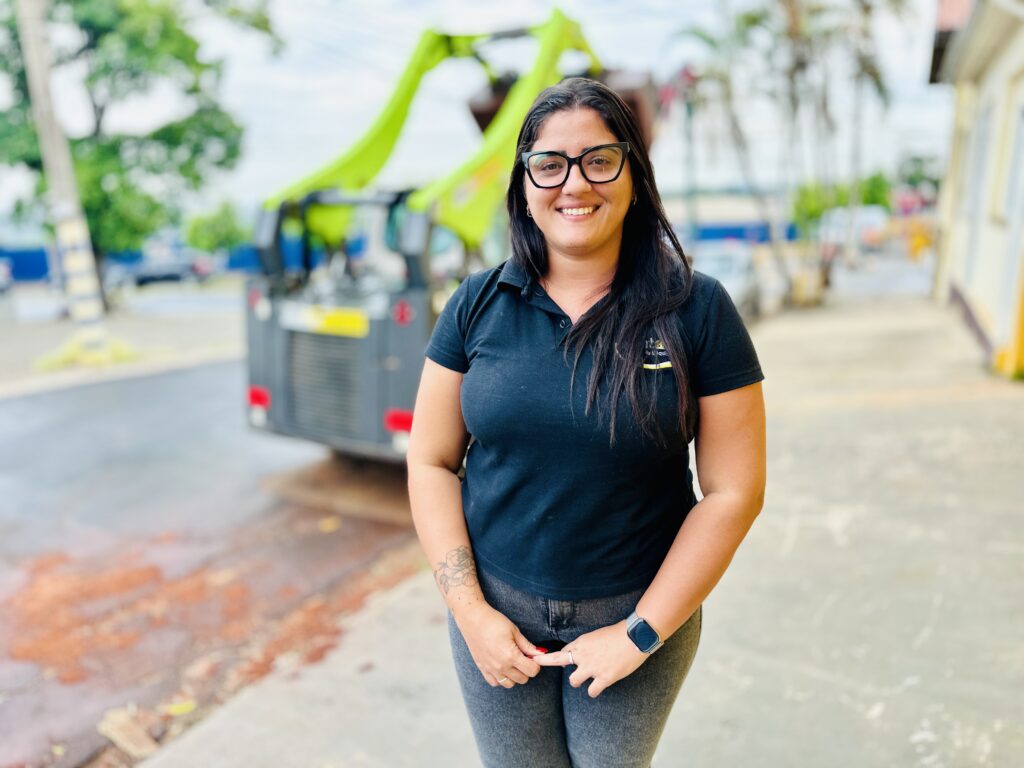 A imagem mostra uma mulher sorridente, posicionada no centro em primeiro plano. Ela tem cabelos longos e escuros, usa óculos de armação preta, uma camisa polo preta e jeans cinza. Em seu braço direito, há uma tatuagem floral visível, e ela está com as unhas pintadas de vermelho. No pulso esquerdo, ela usa um smartwatch. Atrás dela, há uma máquina de construção compacta, do tipo minicarregadeira, pintada em verde-limão e cinza. O fundo está desfocado, mas é possível perceber que a cena ocorre ao ar livre, em uma calçada ou caminho pavimentado próximo a uma estrada, com árvores e um edifício ao longe. A iluminação é clara, sugerindo que a foto foi tirada em um dia nublado ou com luz difusa, e o foco está na mulher, destacando-a em relação ao fundo. A imagem parece ser um retrato profissional, possivelmente relacionado a um ambiente de trabalho na área de construção ou maquinário.