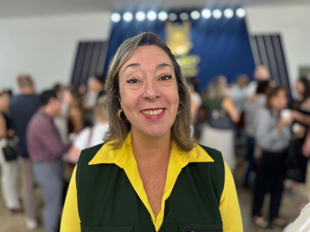 A imagem mostra uma mulher de meia-idade, com cabelo castanho claro, sorrindo de forma acolhedora. Ela está usando uma camisa amarela vibrante com gola e um colete verde escuro por cima. Ela também usa brincos de argola dourados. O fundo está desfocado, mostrando um ambiente interno com várias pessoas ao longe, possivelmente um evento social ou corporativo. Atrás dela, há um painel azul com listras verticais e luzes brilhantes, além de um logotipo dourado desfocado. A iluminação é suave e destaca o rosto da mulher, evidenciando seus traços e batom vermelho. A foto está focada nela, com o fundo desfocado para dar destaque à pessoa.