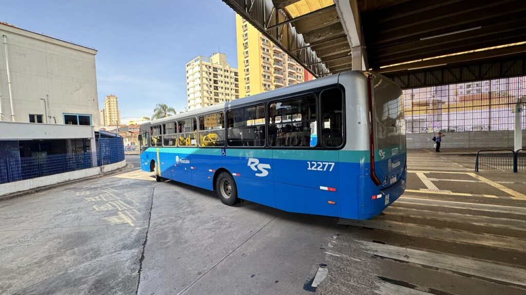 A imagem mostra um ônibus de transporte público em um terminal urbano na cidade de Piracicaba, Brasil. O ônibus é predominantemente azul com detalhes prateados e tem o número de identificação "RS 12277" visível nas laterais e na traseira. A foto foi tirada em um dia ensolarado, com céu azul claro e sombras marcadas sob a cobertura do terminal. Ao fundo, é possível ver prédios residenciais altos, típicos de uma área urbana. Há também uma pessoa ao fundo, próxima à parede do terminal. A perspectiva da foto é ampla, tirada de um ângulo baixo, o que destaca o tamanho do ônibus e a estrutura do terminal. O ambiente transmite a sensação de um local movimentado e funcional para o transporte público da cidade.