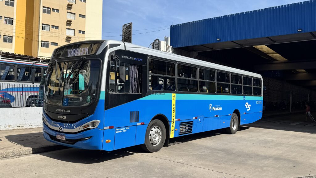 A imagem mostra um ônibus moderno estacionado em um terminal rodoviário. O ônibus é predominantemente azul com detalhes em prata e preto, e possui uma faixa amarela vertical na lateral dianteira onde está escrito "PONTO CEGO". Na parte frontal do veículo, há as marcas "MASCARELLO" e "IVECO", além do modelo "GRANVIA". O número de identificação do ônibus é 57072, e a placa é SWS4G57. Também está visível o selo da Prefeitura de Piracicaba, indicando que o ônibus pertence ao sistema de transporte público dessa cidade. O letreiro eletrônico na frente do ônibus mostra a palavra "GARAGEM", sugerindo que o veículo está fora de operação no momento. O terminal possui uma estrutura metálica azul e, ao fundo, há um prédio residencial amarelo. O céu está claro, indicando que a foto foi tirada durante o dia. O ônibus aparenta estar novo, com fabricação e modelo de 2024.