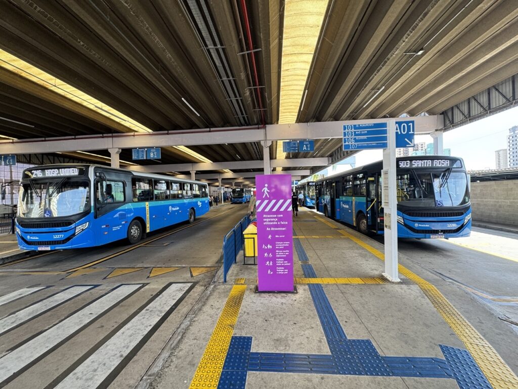 A imagem mostra um terminal rodoviário moderno e espaçoso, provavelmente localizado em Piracicaba, Brasil. O terminal possui um teto alto e curvado com grandes aberturas que permitem a entrada de luz natural, iluminando todo o ambiente. No centro da imagem, há uma faixa tátil azul e amarela para orientação de pessoas com deficiência visual, que se estende ao longo da plataforma de embarque. De ambos os lados da faixa, há dois ônibus grandes e modernos estacionados. O ônibus à esquerda está identificado com o número 246 e o destino "HOSP. UNIMED", enquanto o ônibus à direita é o 503 com destino "SANTA ROSA". Acima das plataformas, placas azuis indicam outras linhas de ônibus, como "101 JD. MONUMENTO", "103 NHÔ QUIM", "105 ALGODÃO", entre outras. No centro da imagem, há uma placa roxa com orientações para os passageiros, destacando regras de segurança como: "Atravesse com segurança utilizando a faixa", "Não corra!", "Crianças sempre acompanhadas dos pais!", "Ofereça auxílio ao idoso e aos deficientes!", além de proibições como andar de skate, patins ou com animais de estimação na área. No fundo, algumas pessoas caminham pela plataforma, e mais ônibus estão estacionados, mostrando que o terminal está em funcionamento e movimentado. A composição transmite uma sensação de organização, segurança e acessibilidade no transporte público local.