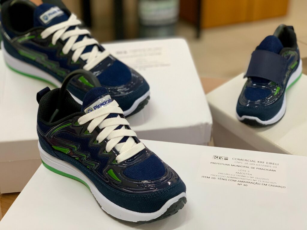 A imagem mostra três pares de tênis esportivos sobre caixas de papelão brancas, provavelmente em um ambiente interno como um escritório ou depósito. Os tênis têm a cor predominante azul-marinho, com detalhes em verde e solado branco. Dois dos tênis são modelos adultos com cadarço branco, enquanto o terceiro tênis, menor, parece ser infantil e possui fechamento em velcro. O tênis em destaque, na frente, está bem focado, enquanto os demais estão desfocados ao fundo. Na caixa onde o tênis está apoiado, há uma etiqueta com informações em português que indicam que se trata de uma amostra para a Prefeitura Municipal de Piracicaba, dentro de um processo licitatório (Pregão Eletrônico Nº 162/2025), com o item descrito como "Tênis com amarração em cadarço", tamanho 30. A imagem transmite a ideia de apresentação ou avaliação de amostras de calçados para um pedido oficial.
