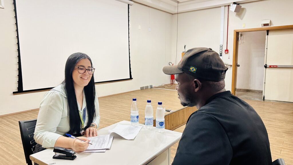 A imagem mostra uma cena em um ambiente interno, possivelmente uma sala de aula, escritório ou auditório. No centro da imagem, há uma mulher sentada atrás de uma mesa branca, sorrindo e aparentemente conversando com um homem que está sentado à sua frente. A mulher tem cabelo longo e escuro, usa óculos e uma blusa verde clara. O homem está de costas para a câmera, usa uma camiseta preta e um boné marrom com uma pequena bandeira do Brasil na lateral. Sobre a mesa, há três garrafas de água da marca "Levity", um grampeador, uma caneta azul e alguns papéis. Ao fundo, uma tela de projeção branca está estendida na parede, e há uma porta com uma placa vermelha escrita "empurre", indicando que a cena provavelmente ocorre no Brasil. A atmosfera parece ser de um atendimento ou entrevista, com a mulher em uma posição de apoio ou orientação, e o homem buscando ajuda ou informação. O ambiente é bem iluminado, com piso de madeira clara e paredes bege.