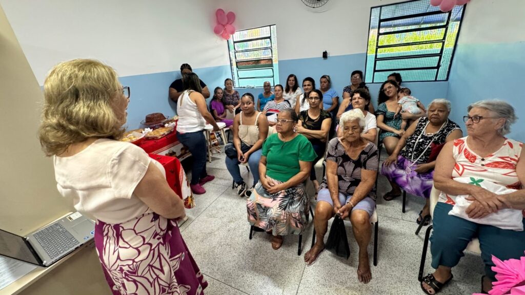 A imagem mostra um encontro em uma sala clara e arejada, com paredes em tons de azul e branco. No centro da cena, uma mulher de cabelos loiros, vestindo uma blusa branca e uma saia com estampa floral roxa, está de costas para a câmera, aparentemente falando ou apresentando algo para um grupo de pessoas sentadas à sua frente. O público é composto majoritariamente por mulheres de várias idades, muitas delas de meia-idade ou idosas, vestindo roupas casuais de verão, como camisetas, regatas e saias estampadas. Algumas delas prestam atenção à palestrante, enquanto outras parecem mais descontraídas. No fundo, uma mulher segura um bebê no colo. À esquerda da palestrante, há uma mesa com um laptop e outra mesa com um pano vermelho que contém alimentos, como pães e um bolo, sugerindo que o evento pode ser social ou comunitário. A sala tem duas janelas com grades metálicas e algumas bexigas cor-de-rosa penduradas no teto, reforçando o clima de confraternização. A iluminação é clara, com luz natural entrando pelas janelas e luz artificial no teto. A atmosfera geral é amigável e informal.
