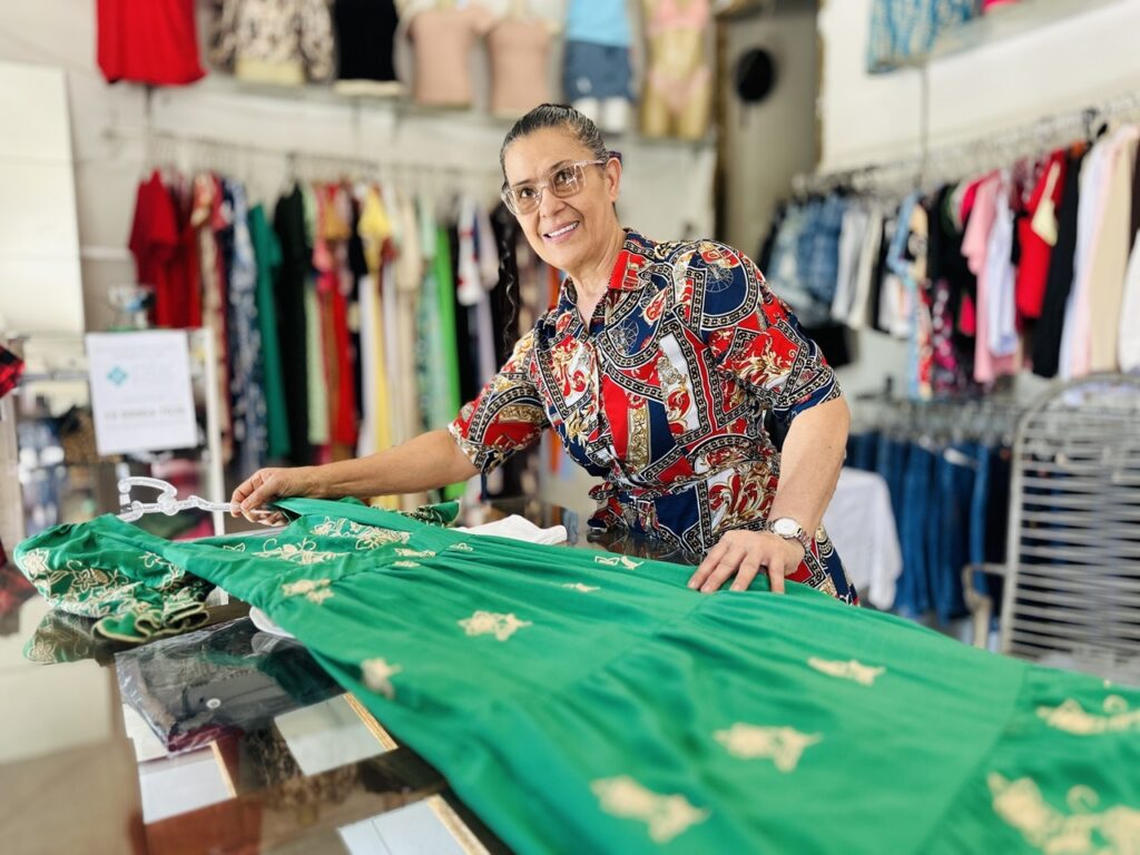 A imagem mostra uma mulher de meia-idade, com cabelos escuros presos para trás e usando óculos, sorrindo de forma acolhedora para a câmera. Ela está atrás de um balcão de vidro, onde está apoiada uma peça de roupa verde com bordados dourados. A mulher veste uma camisa colorida com estampas geométricas e detalhes em vermelho, azul e branco. Ao fundo, há diversas araras com roupas variadas, incluindo vestidos, camisas e calças jeans, além de manequins exibindo outras peças. A iluminação é clara, destacando tanto a mulher quanto a roupa verde em primeiro plano. O ambiente aparenta ser uma loja de roupas, com um clima profissional e ao mesmo tempo convidativo.