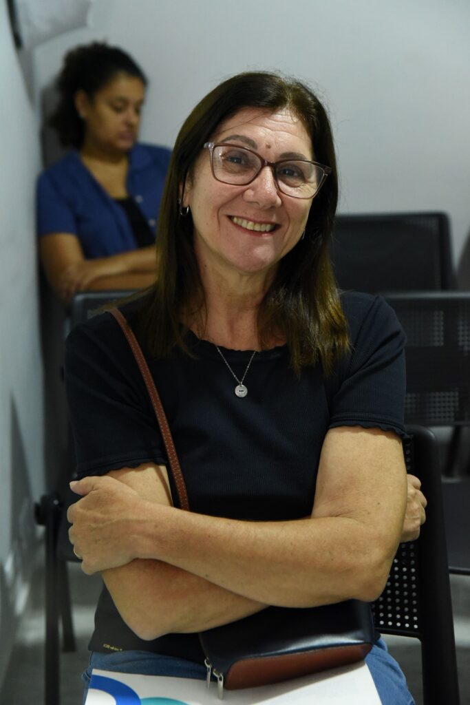 A imagem mostra uma mulher de meia-idade sentada, com os braços cruzados, sorrindo para a câmera. Ela tem cabelos castanhos na altura dos ombros e usa óculos. Está vestindo uma camiseta preta de tecido canelado, jeans azul e um colar prateado com um pingente circular pequeno. Ela carrega uma bolsa de couro marrom pendurada no ombro direito, e no colo segura uma bolsa azul escura. Ao fundo, desfocada, aparece outra mulher com cabelo preso e camisa azul. O ambiente parece ser uma sala de espera ou auditório, com cadeiras pretas de tela. A iluminação é suave, destacando o rosto da mulher principal. Na bolsa azul, há um pequeno texto que diz "K2B with love". A imagem transmite uma sensação de simpatia e conforto.