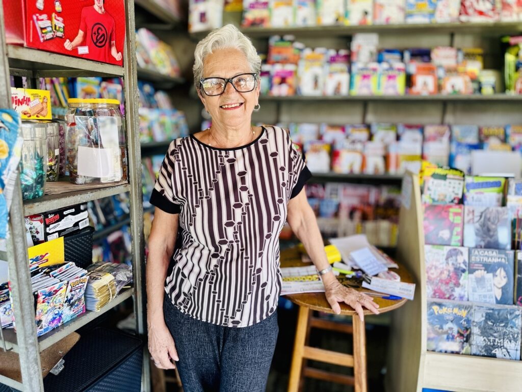 A imagem mostra uma senhora idosa, de cabelos curtos e brancos, usando óculos de armação preta, sorrindo de forma acolhedora para a câmera. Ela está em um ambiente que parece ser uma banca de jornal ou uma pequena loja de hobbies, cercada por várias revistas em quadrinhos, revistas e doces. À sua esquerda, há uma estante metálica cheia de revistas e quadrinhos, incluindo títulos conhecidos como "Pokémon", "Batman" e "X-Men". À direita, há outra estante com mais revistas e produtos. Em frente a ela, há uma mesa pequena com alguns papéis e uma caneta. A iluminação é clara e natural, destacando a figura da senhora e os produtos ao redor. O ambiente transmite uma atmosfera acolhedora e tradicional, típica de uma banca de jornal de bairro.
