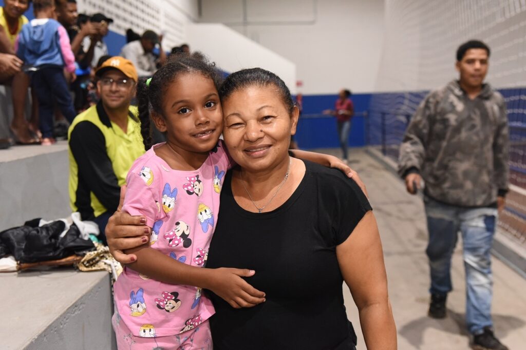 A imagem mostra uma jovem menina e uma mulher mais velha, provavelmente avó e neta, abraçadas e sorrindo para a câmera. A menina tem pele escura, cabelos trançados com detalhes verdes e veste uma camiseta rosa com personagens da Disney (Minnie Mouse e Margarida). A mulher tem pele clara, cabelos escuros presos para trás e veste uma camiseta preta. Elas estão em um ambiente interno, que parece ser uma quadra esportiva ou ginásio, com arquibancadas de concreto ao fundo. Há outras pessoas sentadas e em pé nas arquibancadas, mas o foco principal está na expressão feliz e no abraço carinhoso entre as duas. A iluminação é natural e quente, dando um clima acolhedor à cena.