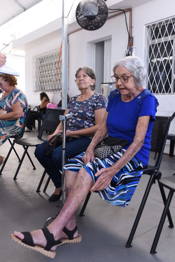 A imagem mostra duas mulheres idosas sentadas em cadeiras de plástico pretas, em um ambiente que parece ser uma sala de espera ou um centro comunitário. A mulher em primeiro plano tem cabelo branco curto, usa óculos, uma camiseta azul vibrante e uma saia listrada azul e branca. Ela apresenta manchas roxas ou hematomas visíveis nos braços e pernas e está olhando para baixo com a boca ligeiramente aberta. Ao lado dela, outra mulher com cabelo grisalho e castanho usa uma camiseta com estampa floral e calça jeans azul escura, olhando para o lado. Entre as duas, há uma bengala prateada apoiada no chão. Ao fundo, há outras pessoas sentadas e elementos do ambiente, como uma parede branca, uma janela com grades, um ventilador preto fixado na parede e uma cobertura branca que deixa a luz natural entrar, indicando que é dia. A cena transmite uma atmosfera de espera e atenção, possivelmente em um local voltado ao atendimento ou apoio a idosos.