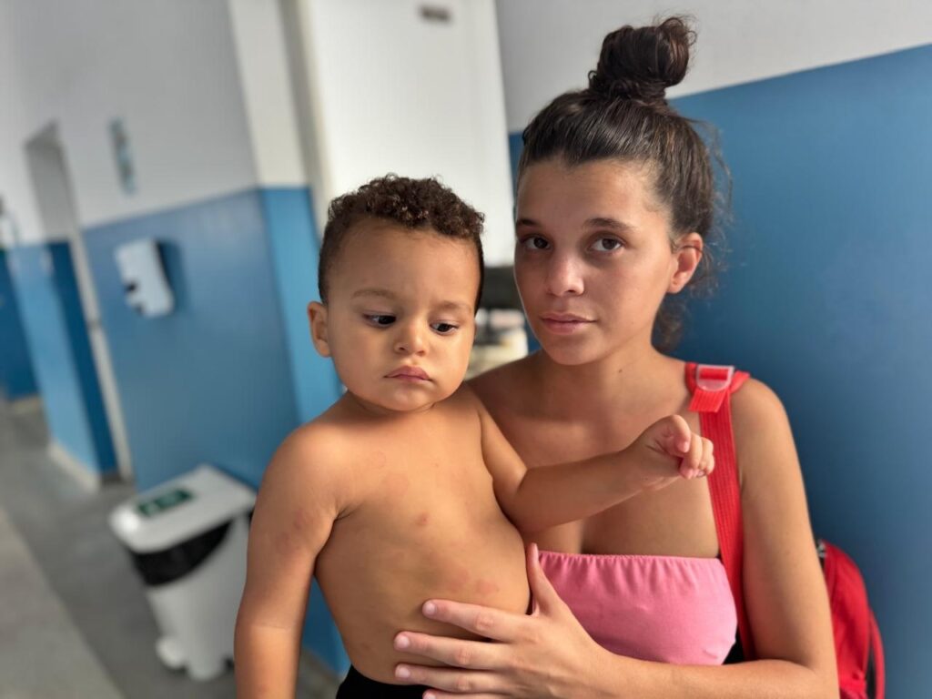 A imagem mostra uma mulher jovem segurando uma criança pequena nos braços, em um ambiente que parece ser um corredor de uma unidade de saúde ou hospital. A mulher tem cabelo escuro preso em um coque alto, veste uma blusa rosa sem alças e carrega uma mochila vermelha. A criança, que está sem camisa, apresenta várias manchas vermelhas arredondadas espalhadas pelo tórax e abdômen, o que pode indicar algum tipo de problema de pele ou doença. Ambos têm expressões sérias ou preocupadas. O fundo é composto por paredes azuis e brancas, um dispenser de álcool em gel e uma lixeira, reforçando a ideia de um ambiente clínico. A iluminação é clara e uniforme, típica de ambientes internos.