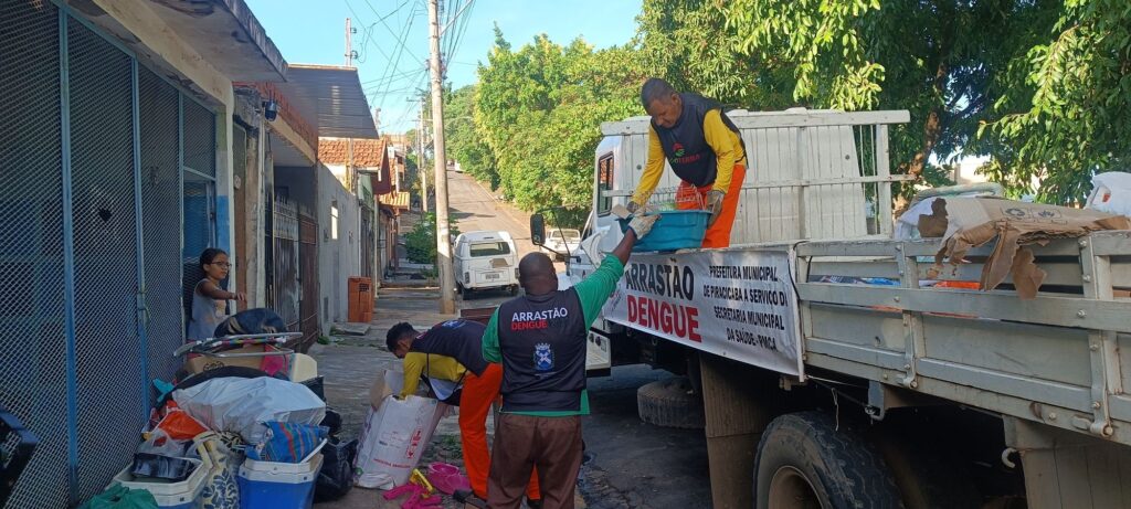 A imagem mostra uma ação comunitária de combate à dengue na cidade de Piracicaba, Brasil, realizada em 2022. Três trabalhadores, vestidos com uniformes azuis, estão recolhendo materiais acumulados na calçada que podem servir como criadouros do mosquito da dengue. Eles estão carregando esses materiais em um caminhão branco, que possui uma faixa com a inscrição "ARRASTÃO DENGUE 2022" e informações da Prefeitura Municipal de Piracicaba e da Secretaria Municipal da Saúde. No canto esquerdo da imagem, uma criança observa a ação de dentro de um portão azul. A rua é inclinada e ladeada por casas simples, com árvores ao fundo e céu azul claro, indicando um dia ensolarado. O cenário transmite a importância do trabalho coletivo e da mobilização para a prevenção da dengue, destacando o esforço da prefeitura para manter a comunidade protegida contra a doença.
