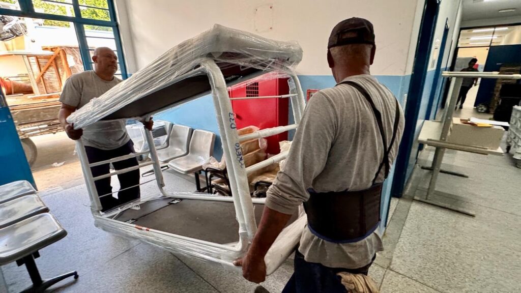 A imagem mostra dois homens carregando manualmente um grande objeto metálico, possivelmente uma estrutura de cama hospitalar ou uma mesa especial, que está envolta em plástico filme transparente para proteção. Eles estão em um corredor interno, provavelmente de um hospital ou outra instituição, dado o ambiente com cadeiras metálicas alinhadas e um armário de incêndio vermelho ao fundo. Um dos homens está de costas, usando boné, camiseta cinza e um cinto de suporte lombar, enquanto o outro está de frente, também com camiseta cinza, demonstrando esforço para mover o objeto pesado. Na estrutura carregada há uma inscrição feita à mão que diz "PRETO D33". A iluminação é clara e uniforme, sugerindo luz natural e artificial. O cenário transmite a ideia de uma movimentação ou instalação de equipamentos novos ou reformados dentro do local.