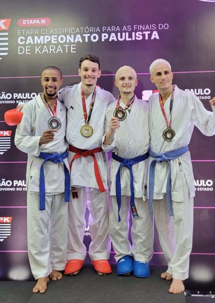 A imagem mostra quatro homens jovens vestidos com quimonos brancos de karatê, posicionados lado a lado em um pódio, celebrando suas conquistas em uma competição. Eles estão em um ambiente interno, com um fundo roxo escuro que traz inscrições em branco e rosa, indicando que o evento é a "ETAPA III" do "Campeonato Paulista de Karatê", realizado em São Paulo, com apoio do governo do estado e da FPK (Federação Paulista de Karatê). Cada competidor usa um cinto colorido (azul ou vermelho) e medalhas penduradas no pescoço, simbolizando suas colocações na competição: o segundo da esquerda usa um cinto vermelho e uma medalha de ouro, enquanto os demais possuem cintos azuis e medalhas de prata ou bronze. Dois deles também usam protetores nos pés e um segura uma luva de combate vermelha. Eles sorriem e aparentam estar felizes, demonstrando espírito esportivo e companheirismo. A iluminação é clara, destacando os atletas contra o fundo escuro.