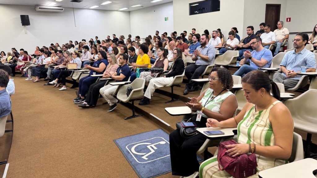 A imagem mostra uma grande audiência sentada em um auditório com assentos em formato de arquibancada. Aproximadamente 60 a 80 pessoas estão presentes, a maioria delas olhando para a frente, provavelmente assistindo a uma apresentação ou palestra. Algumas pessoas no meio estão aplaudindo. As cadeiras são de plástico bege com pequenas mesas acopladas para escrita. O ambiente é interno, com paredes brancas e carpete marrom. No chão, próximo ao primeiro plano da imagem, há um símbolo azul e branco indicando acessibilidade para cadeirantes. A iluminação é clara e uniforme, típica de ambientes fechados para eventos ou aulas. As pessoas estão vestidas de forma casual a social, e o clima geral parece ser de atenção e interesse.