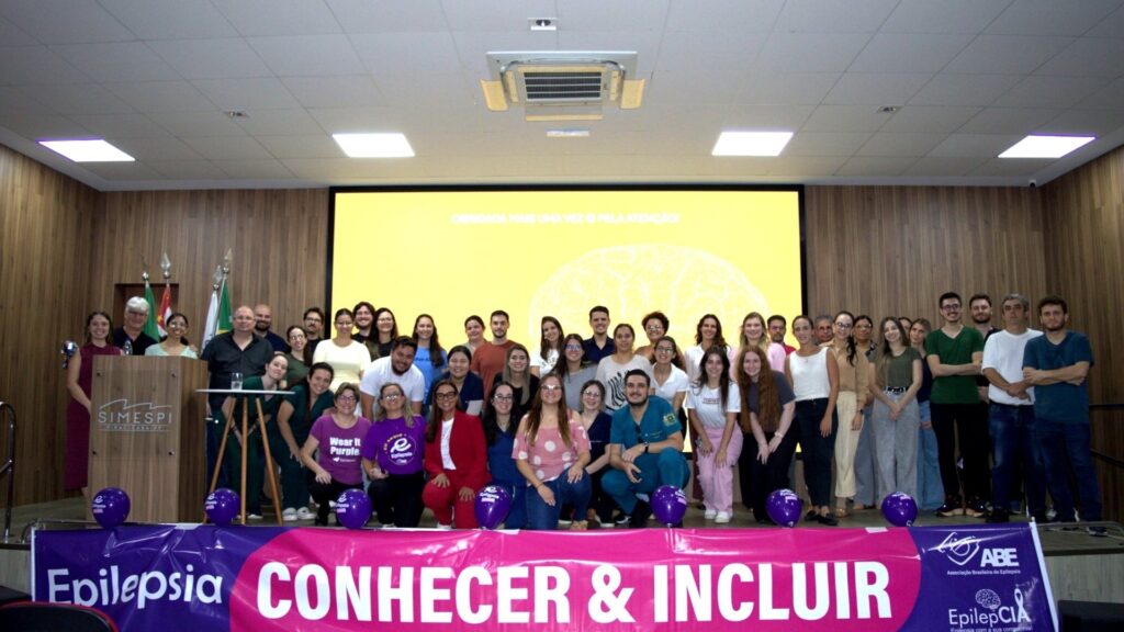 A imagem mostra um grande grupo de pessoas reunidas em um palco, posando para uma foto em grupo. São cerca de 45 indivíduos, entre homens e mulheres de diferentes idades, alguns vestidos com camisetas roxas, outros com roupas casuais e alguns com jalecos médicos, indicando uma possível ligação com a área da saúde. O ambiente é um auditório com paredes de madeira e teto com luminárias embutidas. Ao fundo, há um telão exibindo uma imagem com o desenho de um cérebro e textos em português relacionados à epilepsia, como "Epilepsia CONHECER & INCLUIR" e "EU APOIO Epilepsia". À esquerda do palco, está um púlpito com o logo do SIMESPI (Sindicato dos Médicos de Piracicaba) e algumas bandeiras. Na frente do palco, há um banner roxo e rosa com mais informações e logos, além de balões roxos decorando o espaço. O conjunto da imagem sugere que o grupo está participando de um evento ou campanha de conscientização sobre epilepsia, com foco na inclusão e no apoio às pessoas que convivem com essa condição. A atmosfera transmite união, compromisso e educação em saúde.