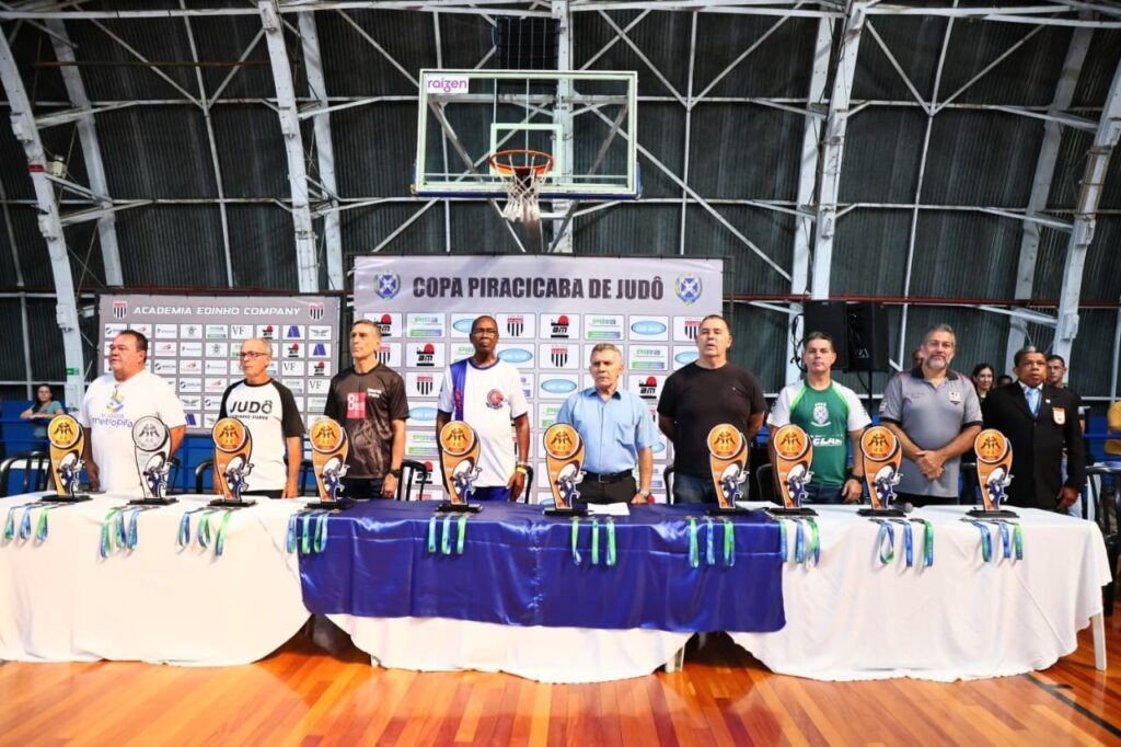 A imagem mostra um grupo de nove homens alinhados atrás de uma mesa longa, dentro de uma quadra esportiva coberta por um telhado alto de estrutura metálica. Eles parecem estar participando de uma cerimônia ou evento oficial, possivelmente relacionado ao judô, conforme indicado pela faixa ao fundo com os dizeres "COPA PIRACICABA DE JUDÔ". Na mesa, coberta por uma toalha branca com um tecido azul por cima, estão expostos vários troféus e muitas medalhas penduradas na frente. Os homens vestem roupas variadas: alguns estão com camisetas esportivas, outros com roupas casuais e um deles está de terno e gravata, sugerindo uma posição de destaque ou autoridade no evento. Ao fundo, além da faixa da copa de judô, há outras faixas com nomes e logotipos de patrocinadores, como "ACADEMIA EDINHO COMPANY" e "raízen". A quadra possui piso de madeira e uma cesta de basquete é visível, indicando que o local é uma ginásio poliesportivo. A iluminação é forte e uniforme, típica de ambientes fechados para eventos esportivos. O clima geral da imagem é de formalidade e celebração esportiva, provavelmente durante a abertura ou premiação de uma competição de judô.