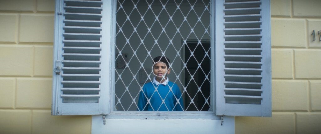 A imagem mostra um menino jovem olhando através de uma janela com grades de metal em formato de losangos, que possui um buraco circular no centro por onde ele está olhando diretamente. A janela tem persianas azuis claras, abertas para os lados, e está instalada em uma parede de cor amarela clara com blocos retangulares. O menino veste uma camiseta azul e uma camisa branca por baixo, e sua expressão é neutra, talvez um pouco curiosa. A luz é suave, sugerindo um dia nublado ou iluminação interna. O número "12" está parcialmente visível na parede à direita da janela. A composição transmite uma sensação de isolamento ou contemplação, com o menino enquadrado exatamente pelo buraco circular da grade.