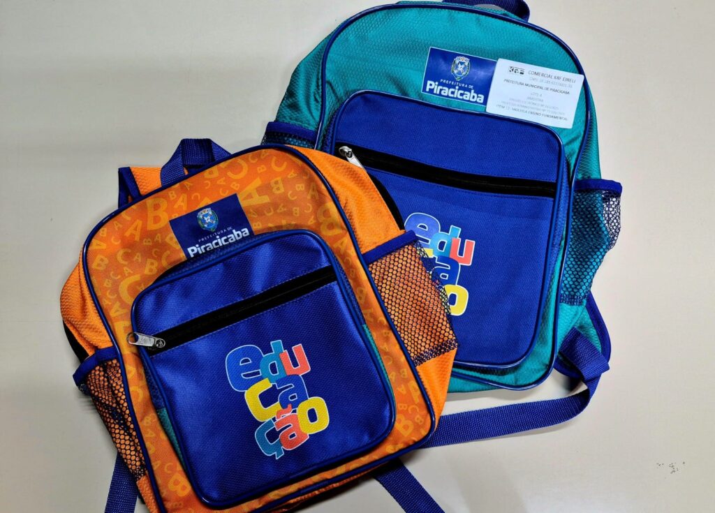 A imagem mostra duas mochilas escolares posicionadas sobre uma superfície clara. A mochila à esquerda é predominantemente laranja com detalhes em azul e apresenta um padrão de letras "A", "B" e "C" espalhadas pelo tecido. Na parte frontal, há um bolso azul com a palavra "Educação" escrita em letras coloridas (azul, vermelho e amarelo). Acima do bolso, está o logotipo da "Prefeitura de Piracicaba". A mochila à direita é verde água com detalhes em azul e possui um bolso frontal azul semelhante ao da mochila laranja, também com o logotipo da "Prefeitura de Piracicaba". Preso na parte superior dessa mochila há uma etiqueta branca com informações da empresa fornecedora, incluindo o nome "COMERCIAL KRF EIRELI", o CNPJ, e dados do processo administrativo da prefeitura. Ambas as mochilas têm bolsos laterais em tela (mesh) e alças ajustáveis na cor azul. A composição da imagem é vista de cima, destacando as mochilas como itens oficiais fornecidos pela prefeitura para o ensino fundamental, conforme indicado na etiqueta.