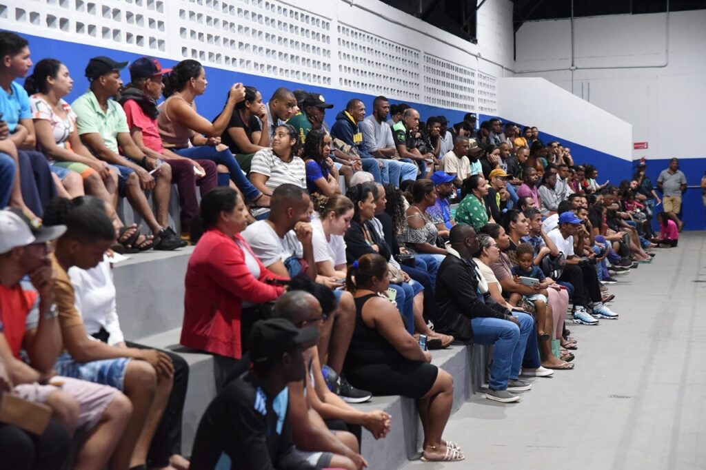 A imagem mostra uma multidão diversificada de pessoas sentadas em arquibancadas de concreto dentro de um ginásio ou centro comunitário coberto. As arquibancadas estão dispostas em níveis, e as pessoas estão sentadas lado a lado, observando algo fora do campo de visão da foto, provavelmente um evento esportivo ou uma reunião pública. A parede ao fundo é branca com uma parte superior que tem um padrão vazado, e a parte inferior é azul. A iluminação é clara e uniforme, típica de ambientes internos. As pessoas vestem roupas variadas, desde camisetas e shorts até jaquetas e bonés, refletindo uma diversidade de idades e estilos. Entre os espectadores, destaca-se uma pessoa usando uma camisa verde com a marca "Crefisa", que é patrocinadora do time Palmeiras, sugerindo um possível interesse esportivo entre o público. A imagem transmite uma sensação de concentração e envolvimento dos presentes com o evento que assistem.