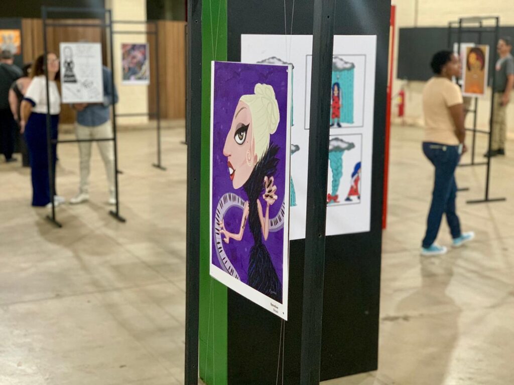 A imagem mostra uma exposição de arte em um ambiente interno, provavelmente uma galeria ou salão. Em destaque, há uma caricatura grande e colorida de Lady Gaga, com traços exagerados típicos desse estilo: cabeça grande, cabelo loiro preso em um coque alto e vestido preto com penas. A caricatura está emoldurada por um fundo roxo e uma representação estilizada de um teclado de piano. A obra está suspensa por fios finos presos a uma estrutura preta e verde. Ao fundo, é possível ver outras obras de arte expostas em painéis semelhantes, além de algumas pessoas observando as peças. Uma mulher vestindo blusa branca e calça escura está próxima a uma das obras, enquanto um homem de camisa azul clara observa outra. Uma mulher de camiseta amarela e calça jeans caminha pelo espaço. O chão é claro e brilhante, e a iluminação é forte e uniforme, típica de um espaço destinado a exposições. No canto inferior direito da caricatura de Lady Gaga, está a assinatura "Synnove Brasil", possivelmente indicando o nome do artista e o país de origem. A imagem transmite uma atmosfera cultural e artística, com foco na arte caricatural e na apreciação do público.