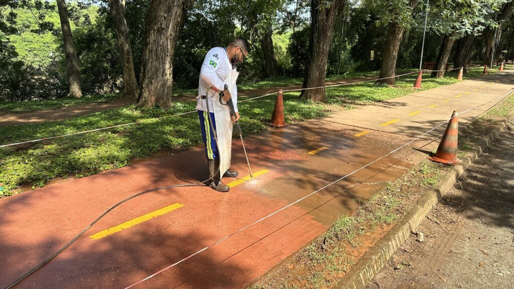 A imagem mostra um homem trabalhando na limpeza de uma ciclovia pavimentada de cor avermelhada, em um parque ao ar livre. Ele está usando um equipamento de alta pressão para lavar o chão. O trabalhador veste uma camisa branca com uma bandeira do Brasil na manga, calça azul com faixas amarelas, avental branco e botas pretas de borracha. O local está cercado por cones laranja e uma corda branca, provavelmente para sinalizar a área de serviço. Ao redor, há árvores grandes e vegetação verde, indicando que é um ambiente natural e tranquilo, iluminado pela luz do dia. A ciclovia segue em diagonal na imagem, e o homem está concentrado na limpeza do caminho.