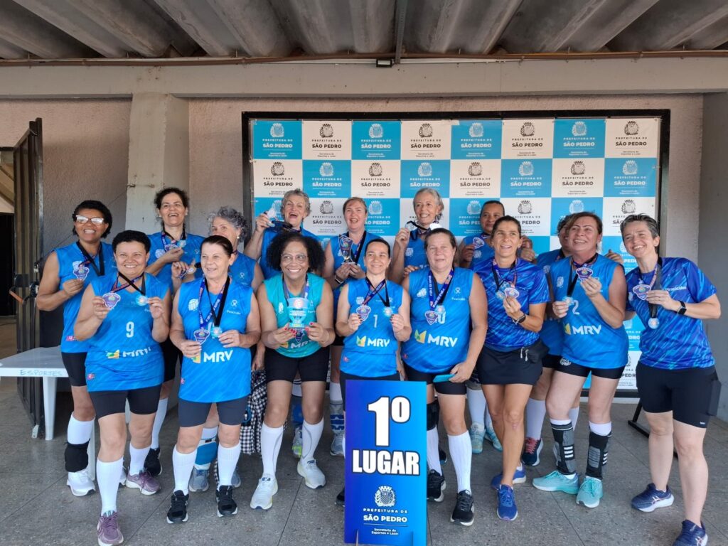 A imagem mostra um grupo de cerca de 14 mulheres, provavelmente uma equipe de voleibol sênior, posando para uma foto comemorativa após uma vitória. Elas estão vestindo uniformes esportivos azuis com o logotipo do patrocinador "MRV" e shorts ou saias pretas. A maioria usa medalhas no pescoço e algumas seguram troféus ou placas. Todas estão sorrindo e aparentam estar felizes com a conquista. Ao fundo, há um banner azul e branco com o logo da "Prefeitura de São Pedro" e a inscrição "Secretaria de Esportes e Lazer". Na frente do grupo, há uma placa azul com a inscrição "1º LUGAR", indicando que a equipe conquistou o primeiro lugar na competição. O ambiente parece ser uma quadra esportiva coberta, com iluminação natural. A diversidade das mulheres é visível, com idades que parecem variar da meia-idade à terceira idade, e diferentes etnias representadas. A imagem transmite um sentimento de alegria, união e conquista esportiva.
