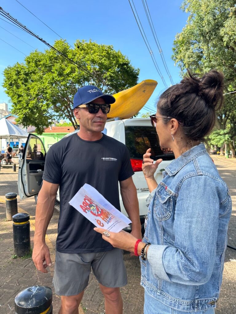 A imagem mostra um homem e uma mulher conversando ao ar livre em um dia ensolarado. O homem, à esquerda, é forte e está vestido com camiseta preta com o logo "SUNTECH", shorts cinza, boné azul com a inscrição "TRC" e óculos escuros. Ele olha para a mulher que está à direita, de perfil, com cabelo preso em um coque, usando jaqueta jeans, óculos escuros e várias pulseiras. Ela segura um folheto branco com textos em vermelho e preto, e parece estar explicando algo, gesticulando com a mão esquerda. Ao fundo, há um SUV branco com um caiaque amarelo preso no teto, e o ambiente parece ser uma área pavimentada próxima a árvores e postes de energia. No chão, há postes baixos pretos, e a iluminação forte indica que é um dia claro e ensolarado. A interação entre os dois parece focada em prevenção, possivelmente relacionada ao conteúdo do folheto que a mulher segura.