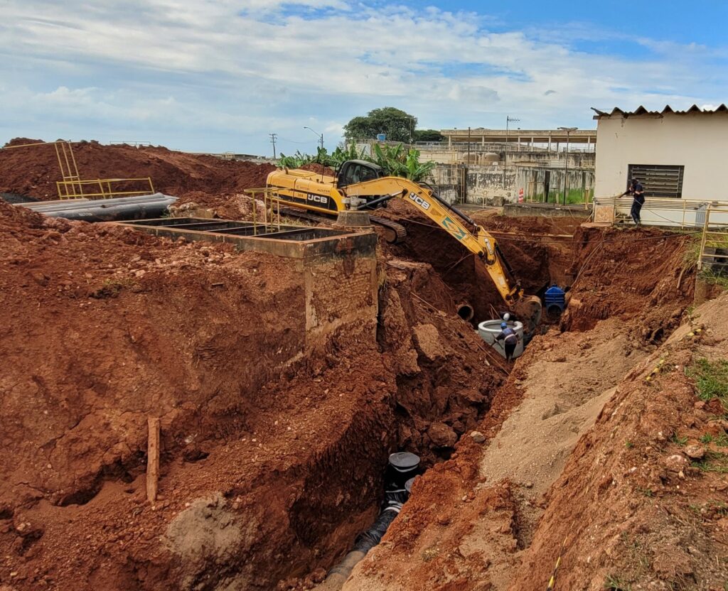 A imagem mostra um canteiro de obras em grande escala, focado em uma escavação profunda para infraestrutura subterrânea. No centro da cena, há uma escavadeira amarela da marca JCB, posicionada em uma plataforma acima de uma vala profunda. O braço da escavadeira está estendido para baixo, dentro da vala, onde um trabalhador usando capacete azul e colete refletivo está próximo a um anel ou segmento de concreto. O solo é de cor avermelhada, típica de terra argilosa, e está acumulado em grandes montes ao redor da escavação. Na parte inferior da vala, há um tubo preto com uma tampa vertical. À esquerda, vários tubos escuros estão empilhados no chão, e uma estrutura de concreto com corrimões metálicos está parcialmente enterrada. Ao fundo, há um prédio industrial branco com telhado de chapas onduladas, um homem em uma plataforma e vegetação verde, incluindo bananeiras. O céu está claro, com algumas nuvens brancas, indicando que a foto foi tirada durante o dia. A imagem transmite a dimensão do trabalho de terraplenagem em relação às máquinas e aos trabalhadores presentes.