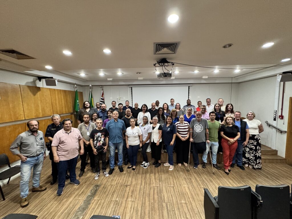 A imagem mostra um grupo grande de pessoas reunidas em uma sala, posando para uma foto em grupo. Elas estão organizadas em três fileiras: algumas pessoas estão de pé no chão, outras estão em um pequeno palco elevado. O grupo é diversificado, com homens e mulheres de diferentes idades, e todos vestem roupas casuais ou sociais. Um homem na primeira fila está usando muletas. A sala tem paredes de madeira à esquerda e ao fundo há uma tela de projeção branca. À esquerda, há três bandeiras: uma verde, uma branca com detalhes verdes e a bandeira do estado de São Paulo. O teto tem luzes embutidas que iluminam bem o ambiente, e há um projetor pendurado no centro do teto. No primeiro plano da imagem, é possível ver a parte superior de cadeiras pretas, sugerindo que a foto foi tirada em um auditório ou sala de conferências. O ambiente transmite a sensação de uma reunião formal, como um seminário, workshop ou evento institucional.