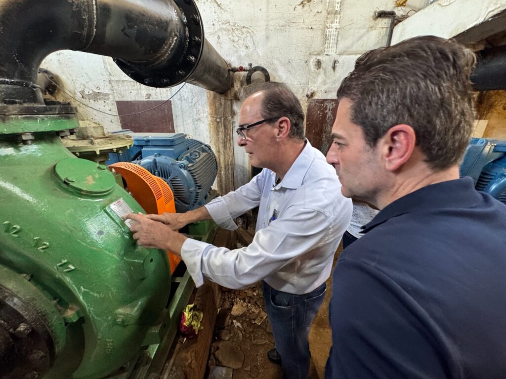 A imagem mostra dois homens em um ambiente industrial, possivelmente uma sala de bombas ou uma casa de máquinas. O homem à esquerda, de meia-idade, usa óculos, uma camisa azul clara e calça jeans. Ele está apontando para uma placa de identificação metálica fixada em uma grande bomba industrial verde. O homem à direita, mais jovem, veste uma camisa polo azul escura e observa atentamente o local indicado. O ambiente é simples e um pouco desgastado, com paredes de concreto pintadas de branco, onde a tinta está descascando em alguns pontos. O chão parece sujo, típico de um local industrial. A bomba verde é grande e robusta, conectada a tubulações pretas grossas por flanges aparafusadas. Ao lado da bomba há um motor azul com uma proteção laranja. A iluminação é artificial, provavelmente fluorescente, e vem de cima, criando sombras suaves. O foco da imagem é a interação entre os dois homens e o equipamento, sugerindo uma inspeção, manutenção ou explicação técnica. Na bomba, está gravado o número "12 12 17".