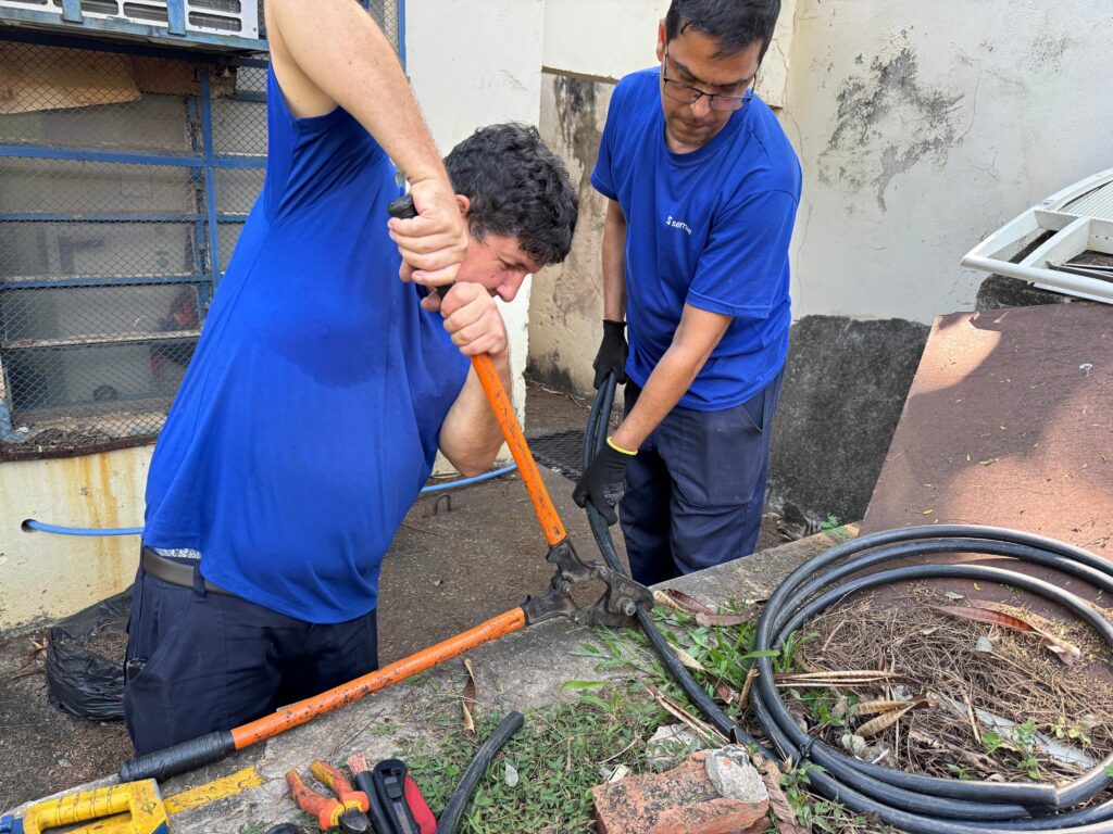 A imagem mostra dois homens trabalhando ao ar livre, provavelmente em um serviço relacionado a cabos elétricos pesados. O homem à esquerda está usando um alicate de corte grande, com cabo laranja, para cortar um cabo preto grosso, mostrando esforço e concentração. Ele veste uma camiseta azul clara que está visivelmente suada e calça escura. O outro homem, à direita, também veste camiseta azul clara com o logo "semae" e calça escura, está segurando o cabo firme para ajudar no corte, usando luvas de proteção. Eles estão em um ambiente externo, com uma parede branca e uma janela gradeada azul ao fundo. No chão próximo a eles, há uma bobina grande de cabo preto, algumas ferramentas espalhadas, como um alicate, uma faca e um nível amarelo, além de folhas secas e grama rala. A luz é forte, indicando que a foto foi tirada durante o dia. A imagem transmite a ideia de trabalho físico e cooperação entre os dois homens para realizar a tarefa.