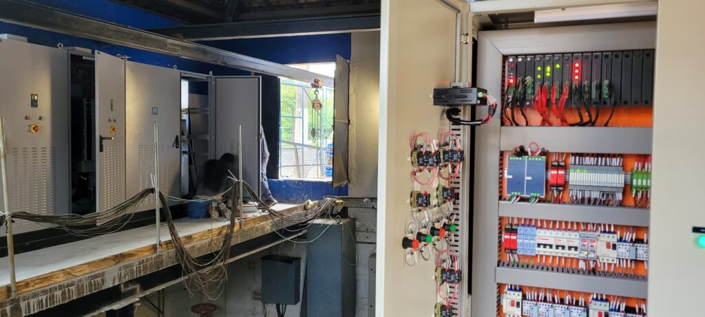 A imagem mostra um ambiente industrial, provavelmente uma sala de controle ou subestação elétrica, onde está sendo realizada manutenção ou instalação. À esquerda, há vários painéis elétricos grandes e cinza, com ventilação e botões de controle. Um técnico está agachado, trabalhando na fiação de um desses painéis. À direita, há um painel elétrico aberto, exibindo diversos componentes eletrônicos, como controladores lógicos programáveis (CLPs) da Siemens com luzes indicadoras vermelhas e verdes, blocos terminais, disjuntores e uma grande quantidade de fios organizados em canaletas cinzas. Na porta do painel, há interruptores e indicadores montados. Ao fundo, é possível ver uma janela ou porta aberta, por onde entra luz natural, iluminando parte do ambiente e revelando árvores e uma cerca do lado de fora. A cena transmite a ideia de um trabalho técnico detalhado em andamento, com boa organização dos cabos e equipamentos.