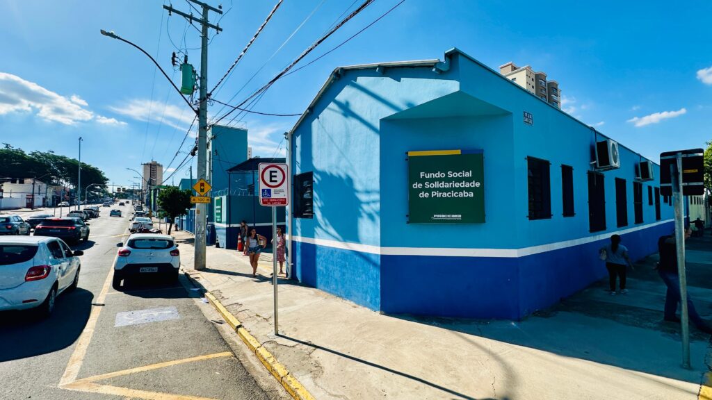 A imagem mostra um prédio pintado em tons de azul localizado em uma esquina da Rua Santa Cruz, em Piracicaba, Brasil. O prédio pertence ao "Fundo Social de Solidariedade de Piracicaba", conforme indicado por uma placa verde grande na fachada. A parte superior do prédio é pintada de azul claro, enquanto a base é azul mais escuro, separadas por uma faixa branca horizontal. Em frente ao prédio, há uma calçada onde duas mulheres estão caminhando próximas à entrada, e outra pessoa caminha afastada, pela lateral do prédio. Ao lado esquerdo da imagem, uma rua movimentada com vários carros estacionados e em movimento se estende até o horizonte, sob um céu azul claro com poucas nuvens. No canto da imagem, há um poste de energia com transformador verde e vários fios aparentes. Placas de sinalização indicam que a vaga é exclusiva para deficientes físicos, com uso obrigatório do cartão, e há uma placa de trânsito de "proibido estacionar". A iluminação é intensa e natural, típica de um dia ensolarado. A composição da foto utiliza a esquina do prédio como ponto de convergência, criando profundidade visual ao longo da rua e da lateral do edifício.