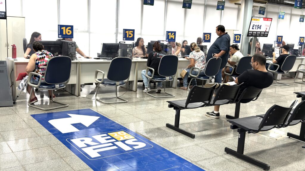 A imagem mostra um ambiente interno de atendimento público, possivelmente uma repartição pública ou setor de serviços da prefeitura de Piracicaba, no Brasil. Há vários balcões numerados de 10 a 16, onde servidores atendem pessoas sentadas em cadeiras. Alguns atendimentos são específicos para "Dívida Ativa", conforme indicado nos balcões 10 a 13. No chão, há uma grande seta azul com o texto "REFIS 2026", indicando um programa de refinanciamento de dívidas. O local é bem iluminado, com teto de estrutura metálica e piso de cerâmica cinza brilhante. No primeiro plano, há uma fileira de cadeiras pretas onde algumas pessoas estão sentadas aguardando atendimento. Uma delas está olhando para o celular. Os atendentes trabalham em computadores atrás dos balcões, protegidos por divisórias transparentes. Acima dos balcões, há painéis digitais exibindo números de senhas para organização do atendimento, como "E194". No geral, o ambiente transmite uma sensação de organização e funcionalidade, com uma clara divisão entre a área de espera e a área de atendimento. A presença do logo da prefeitura e da Fatec Piracicaba sugere que o local está vinculado a órgãos públicos locais e talvez a uma instituição de ensino.