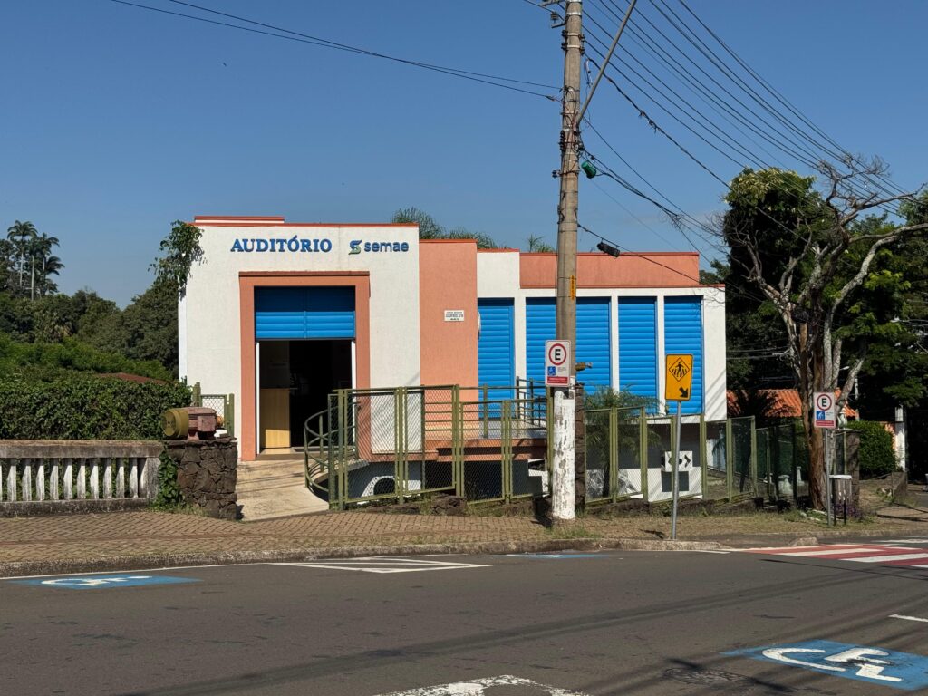 A imagem mostra a fachada de um auditório identificado como pertencente ao SEMAE Piracicaba. O prédio tem arquitetura simples, com paredes em tons de branco e salmão, e portas ou painéis azuis metálicos. Acima da entrada principal está escrito “AUDITÓRIO” junto ao logotipo do Semae. Na parte frontal, há uma rampa de acessibilidade com corrimão, cercada por uma grade metálica verde. Em frente ao local, passa uma rua asfaltada com sinalização horizontal, incluindo vaga para pessoas com deficiência. Também há placas de trânsito, como indicação de proibido estacionar e sinalização de travessia de pedestres.