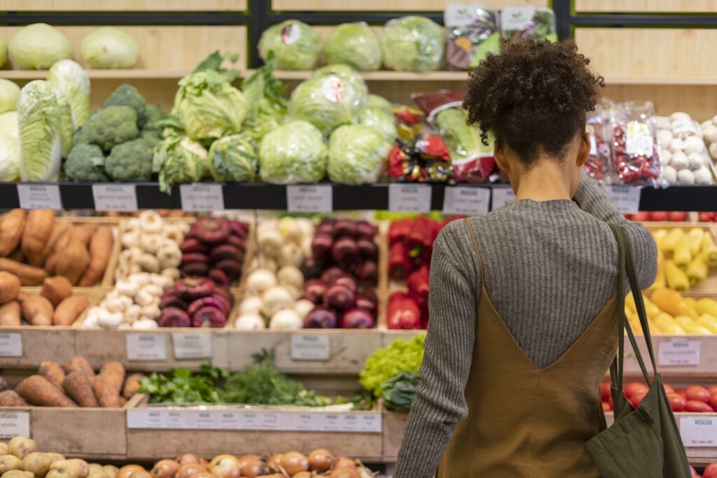 A imagem mostra uma mulher de cabelos cacheados, vista de costas, fazendo compras na seção de hortifrúti de um supermercado. Ela veste uma blusa cinza de manga longa e um vestido tipo jardineira marrom por cima, além de carregar uma bolsa verde escura no ombro direito. À sua frente, há várias prateleiras organizadas com diferentes tipos de legumes e verduras frescas, como repolho, brócolis, alface, pimentões vermelhos, cogumelos, batata-doce, alho, cebolas e cenouras. A iluminação é clara, destacando as cores vibrantes dos alimentos. A foto transmite uma sensação tranquila e cotidiana, típica de uma ida ao supermercado.