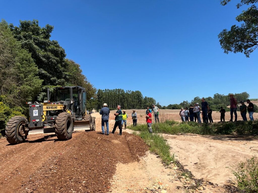 A imagem mostra uma cena ao ar livre em um dia ensolarado com céu azul claro. No centro da imagem, há uma máquina de terraplanagem da marca New Holland, de cor amarela e preta, posicionada em uma estrada de terra. A máquina está com a lâmina voltada para o chão, aparentemente em operação ou pronta para trabalhar. À direita da imagem, um grupo de pessoas, em torno de 15 a 20 indivíduos, está reunido na beira da estrada, algumas em um pequeno barranco coberto de grama. Eles parecem estar observando a máquina ou participando de uma reunião ou evento relacionado à obra. As pessoas vestem roupas variadas, desde roupas casuais até coletes de alta visibilidade, indicando que alguns podem estar envolvidos na obra ou fiscalização. Ao fundo, há uma área rural com árvores verdes, incluindo uma fileira de eucaliptos, e um campo aberto com solo avermelhado. A luz do sol é forte, criando sombras marcadas e destacando a poeira do terreno e os detalhes das rodas da máquina. A cena transmite uma interação entre a comunidade local e o trabalho de manutenção ou construção da estrada em um ambiente rural.
