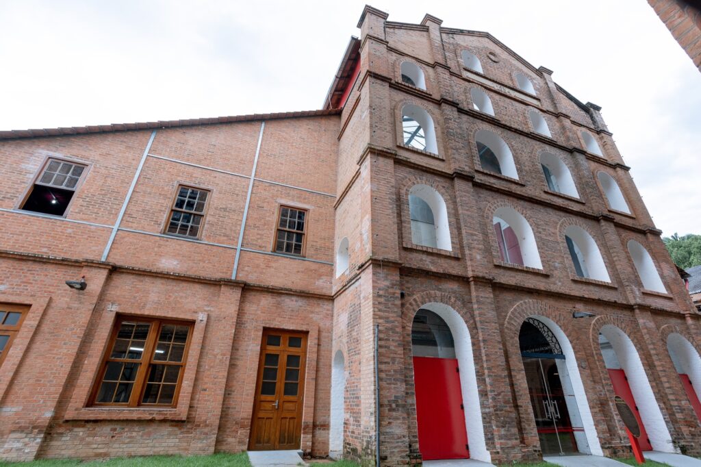 A imagem mostra um edifício histórico de tijolos vermelhos, que parece ser um teatro municipal, conforme o texto extraído. O prédio é grande, com duas seções principais: à direita, uma parte mais alta com vários andares e janelas em arco, algumas com vidros e outras abertas; à esquerda, uma ala mais baixa, com janelas retangulares e uma porta de madeira. A fachada tem muitos detalhes em tijolos, com um estilo arquitetônico antigo, provavelmente industrial ou colonial. A parte mais alta tem entradas amplas em arco no térreo, uma delas com uma porta vermelha e outra com uma porta de vidro, indicando que o edifício pode ter sido reformado para uso moderno. O céu está nublado, com luz difusa, e há um pequeno gramado na frente do prédio. A foto foi tirada de um ângulo baixo, realçando a altura e imponência da construção. A imagem transmite um senso de história e cultura, sugerindo que o local é um ponto importante da cidade.