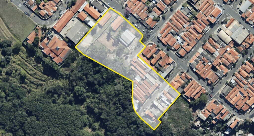 A imagem mostra uma vista aérea de uma área urbana ao lado de uma floresta densa. No centro da imagem, há um terreno grande e irregular demarcado por uma linha amarela, que está parcialmente coberto por uma camada branca translúcida. Dentro desse terreno, há algumas construções, incluindo um edifício maior com telhado branco e várias construções menores com telhados laranja, além de áreas verdes com árvores. Ao redor desse terreno, na parte superior e à direita da imagem, há um bairro residencial com casas próximas umas das outras, todas com telhados laranja característicos, organizadas em ruas estreitas e retas. Na parte inferior e à esquerda da imagem, há uma floresta densa, com árvores de copa fechada, que contrastam com a área urbana. A iluminação indica que a foto foi tirada em um dia claro, com o sol alto no céu, pois as sombras são curtas e definidas. A imagem parece ser utilizada para planejamento urbano ou análise imobiliária, dada a clareza e o destaque do terreno demarcado.