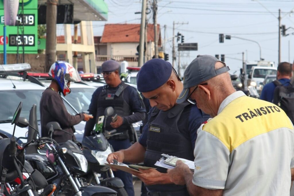 A imagem mostra uma abordagem de trânsito realizada por agentes e policiais em uma rua urbana durante o dia. No primeiro plano, um agente de trânsito, identificado pela camisa branca com detalhes amarelos e a palavra "TRÂNSITO" nas costas, está conferindo documentos junto a um policial fardado com colete tático azul e boina azul. Ao fundo, outro policial, também com uniforme azul escuro e boina, conversa com um motociclista que usa capacete colorido e camisa marrom. Há várias motocicletas estacionadas próximas, além de carros oficiais da polícia. Ao fundo, é possível ver um posto de combustível com os preços dos combustíveis exibidos em painéis verdes. A cena transmite uma ação de fiscalização de trânsito em um ambiente urbano, com iluminação natural e clima claro.