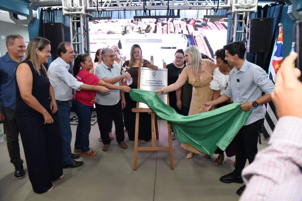 A imagem mostra uma cerimônia de inauguração, onde um grupo de cerca de onze pessoas está reunido ao redor de um cavalete de madeira. No centro, uma mulher vestindo um vestido bege claro e um homem de camisa azul clara estão puxando um tecido verde para revelar uma placa prateada. A placa indica o nome da escola municipal: "Escola Municipal Professora Márcia Regina Giuliani Novello". As pessoas ao redor sorriem e observam o momento da revelação com interesse e satisfação. Ao fundo, há uma estrutura metálica que sustenta uma tela grande mostrando uma imagem aérea de um prédio com painéis solares no telhado, provavelmente a escola que está sendo inaugurada. Também é possível ver a bandeira do estado de São Paulo à direita da imagem. O ambiente parece ser interno, bem iluminado, e o clima é de celebração e formalidade.