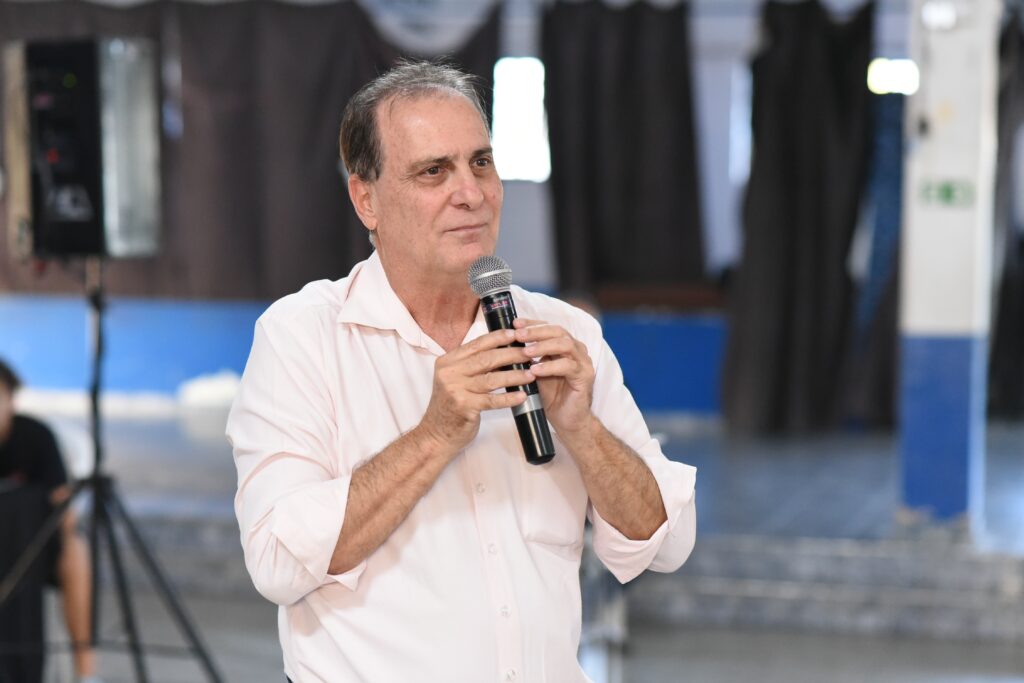 A imagem mostra um homem de meia-idade, com cabelo curto e pele clara, usando uma camisa social rosa claro. Ele está segurando um microfone preto da marca Shure, modelo SM58, com ambas as mãos próximo ao peito. O homem parece estar falando ou se preparando para falar, com uma expressão facial serena e um leve sorriso, olhando para o lado direito da imagem. O fundo está desfocado, mas é possível distinguir um ambiente interno, possivelmente uma sala de eventos ou auditório, com pilares pintados de azul e branco e uma caixa de som sobre um tripé à esquerda. A iluminação é suave e natural, destacando o rosto do homem. A composição transmite a sensação de uma apresentação ou discurso em um evento formal ou comunitário.