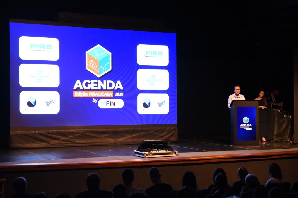 A imagem mostra um evento profissional em um auditório escuro, com um palco iluminado. À esquerda, há uma grande tela digital azul exibindo o texto "AGENDA Edição: PIRACICABA 2026 by PiN", junto com um logotipo em forma de cubo 3D e ícones relacionados a crescimento e moradia. Na tela também aparecem os logos dos patrocinadores, como PIRA, SEBRAE e EPTV. À direita da tela, um homem vestido com uma camisa clara está falando em um púlpito azul escuro, que também tem a marca do evento "AGENDA". Ao lado dele, uma mulher de vestido escuro está em pé, observando. Na frente do palco, é possível ver a silhueta da plateia sentada, assistindo à apresentação. A iluminação destaca o palco, criando um contraste com o restante do ambiente, típico de conferências ou palestras. A foto é tirada do ponto de vista da audiência, mostrando a dimensão do evento e a tela com as informações principais.