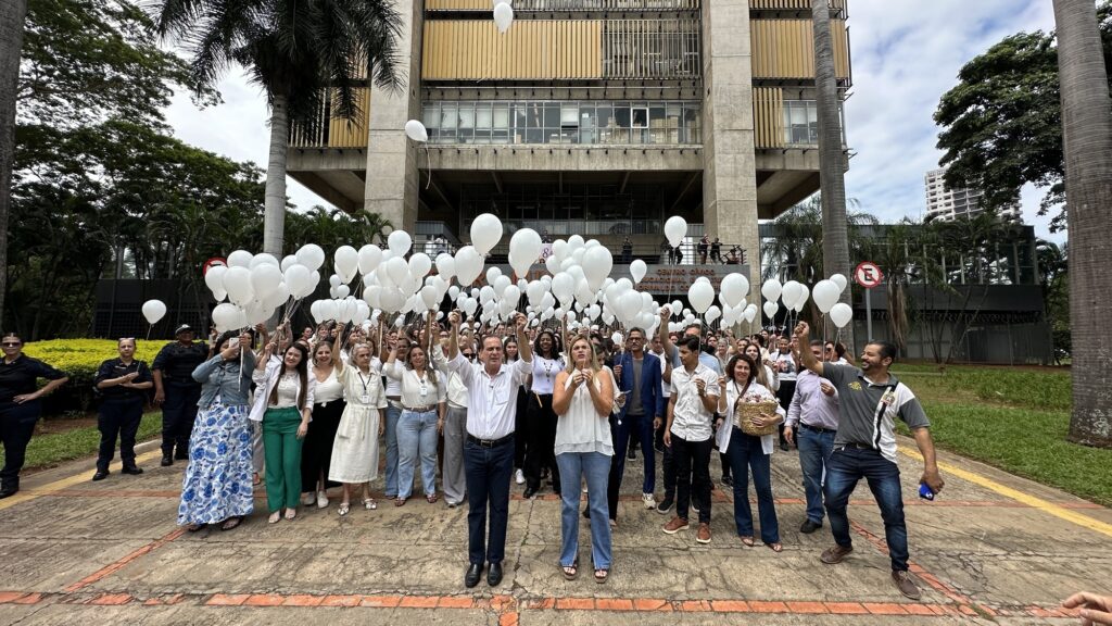 A imagem mostra uma grande reunião de pessoas em um espaço aberto, provavelmente um pátio ou praça, em frente a um edifício de arquitetura brutalista com pilares de concreto e janelas com persianas verticais amarelas. A maioria dos presentes está vestida de branco ou cores claras, e muitos seguram balões brancos, alguns já soltos no ar. No centro da imagem, um homem e uma mulher, ambos com roupas claras, parecem liderar o evento. À esquerda, há algumas pessoas com uniformes, possivelmente policiais ou seguranças. O ambiente é arborizado, com árvores verdes e palmeiras ao redor. A atmosfera sugere um evento solene, como uma homenagem ou memorial, realizado durante o dia com luz natural difusa. O edifício ao fundo é identificado parcialmente por um letreiro que diz "CENTRO CÍVICO EDUCACIONAL E CULTURAL Florivaldo Coelho Prates". A cena transmite um sentimento de união e respeito.