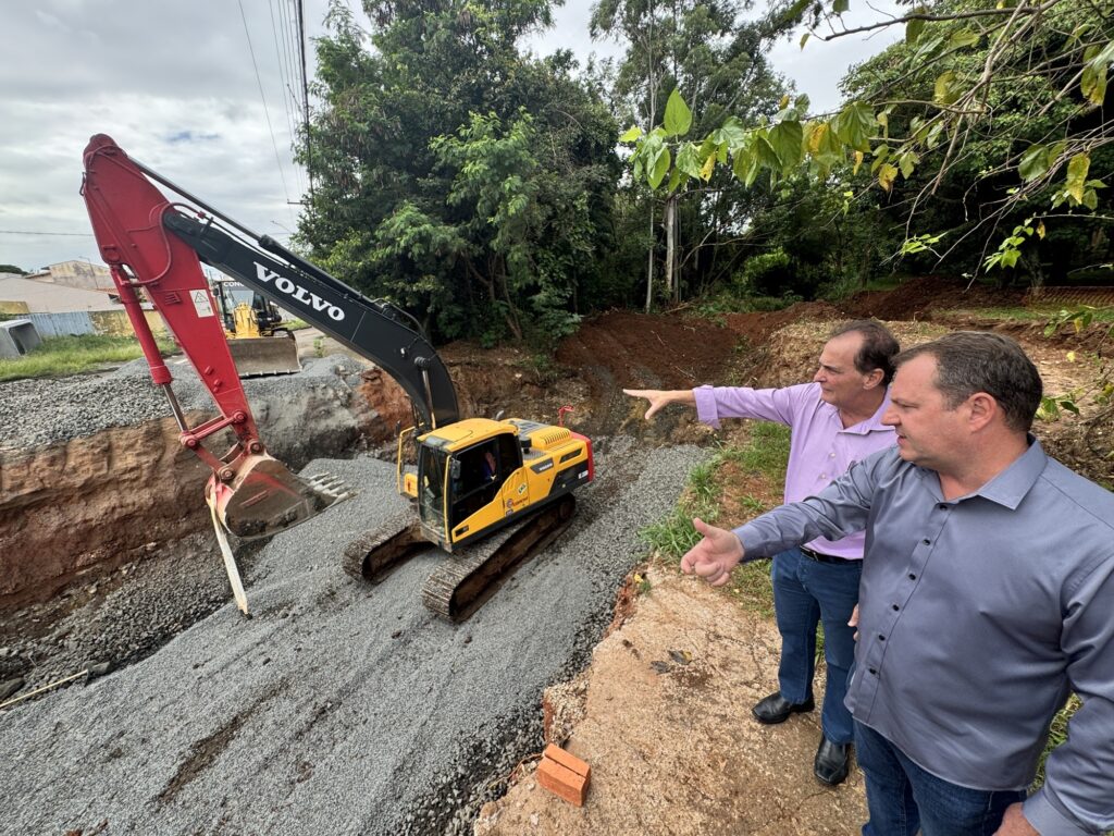 A imagem mostra um canteiro de obras ao ar livre, onde duas pessoas estão em primeiro plano observando e apontando para uma escavadeira amarela da marca Volvo, que está operando em uma vala preenchida com cascalho cinza. Um dos homens, à esquerda, veste uma camisa lilás clara e calça jeans azul, enquanto o outro, à direita, usa uma camisa cinza e também calça jeans. Eles parecem estar discutindo ou analisando o trabalho que está sendo realizado. Ao fundo, há outra máquina amarela e uma área de vegetação com árvores verdes. O céu está nublado, o que proporciona uma iluminação difusa e suave na cena. O chão onde os homens estão é uma mistura de terra e pavimento quebrado. A escavadeira está com a lança levantada, destacando-se na imagem pelo seu tamanho e cor vibrante.