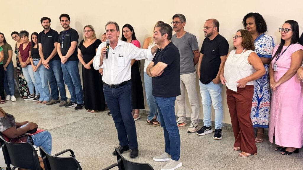 Na imagem aparecem um senhor de camisa branca, calça jeans e sapatos pretos, segurando o microfone na mão direita e estende a mão esquerda num senhor ao seu lado, que veste camiseta preta e calça jeans. Atrás aparecem enfileirados homens e mulheres, prestando atenção na fala do senhor de camisa branca.