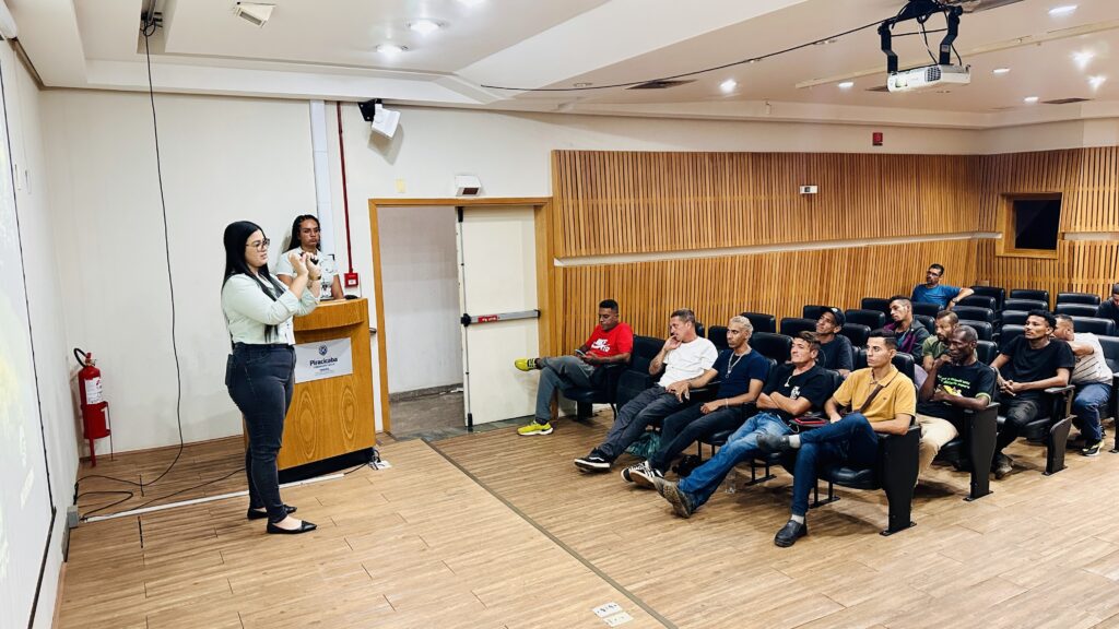 A imagem mostra uma sala de auditório onde uma mulher está fazendo uma apresentação para um grupo de homens sentados em cadeiras. Ela veste uma blusa verde clara e calça escura, e está em pé próximo a um púlpito de madeira que tem a inscrição "Piracicaba TRABALHO SÉRIO CIDADE". Ao fundo, outra mulher está atrás do púlpito, aparentemente acompanhando a apresentação. A plateia é composta principalmente por homens, alguns usando camisetas coloridas, como uma vermelha com o logo "NIKE AIR". A sala tem paredes de madeira clara e teto branco com iluminação embutida. Há uma placa de saída ("SAÍDA") acima da porta, indicando que o local provavelmente fica no Brasil. Também é possível ver um extintor de incêndio na parede. O ambiente sugere uma reunião formal ou uma palestra em um espaço público ou corporativo, com foco em trabalho ou treinamento.