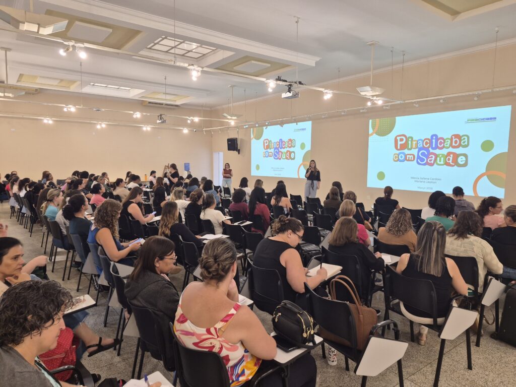 A imagem mostra uma grande sala de conferências ou auditório onde está acontecendo um seminário ou apresentação intitulada "Piracicaba com Saúde". O ambiente é bem iluminado, com paredes claras e iluminação branca no teto. Na frente da sala, duas telas de projeção exibem o título do evento, os nomes das apresentadoras Márcia Juliana Cardoso e Mariana Liepkan, além da data "Março 2024". Também aparece o logo da Prefeitura de Piracicaba. O público é composto majoritariamente por mulheres, sentadas em cadeiras com pequenas pranchetas acopladas, algumas anotando ou olhando para seus celulares. A palestrante está em pé, no centro da frente da sala, entre as duas telas, falando para os participantes. O ambiente transmite uma atmosfera profissional e organizada, sugerindo que o evento é uma iniciativa de saúde pública promovida pela prefeitura local.